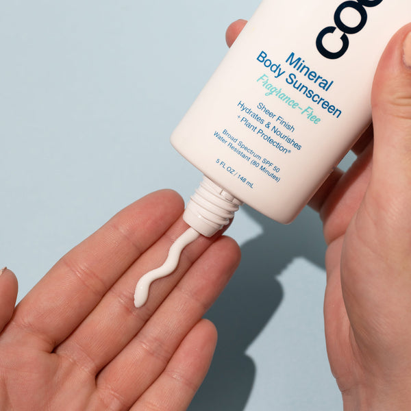 Close-up of a hand dispensing light lotion from a tube of Mineral Body Organic Sunscreen Lotion SPF 50 - Fragrance Free | COOLA on a light blue background, highlighting skincare and body care.