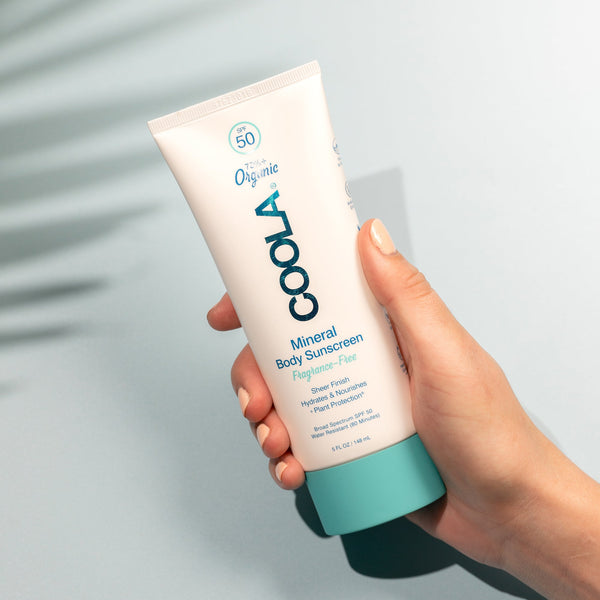 Hand holding a tube of Mineral Body Organic Sunscreen Lotion SPF 50 - Fragrance Free | COOLA against a light background, showcasing the product designed for skincare and body care.