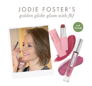 Jodie Foster smiling while applying makeup, alongside a display of Vitamin Glaze® Oil Infused Lip Gloss – Violet Orchid | Farmhouse Fresh with pink and violet lip colors shown.