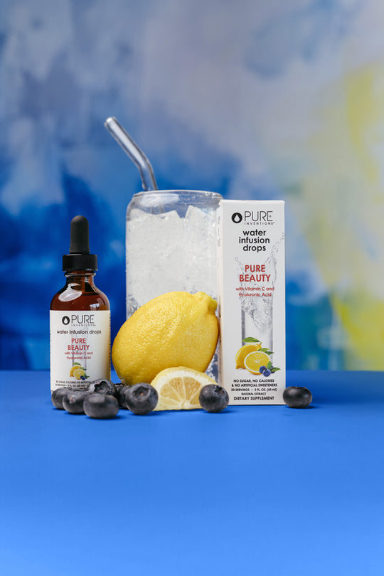 Bottle and box of Pure Beauty Water Infusion Drops - Berry Lemonade | Pure Inventions displayed with a glass of ice, lemon, and blueberries on a blue surface against a blurred blue and yellow background.