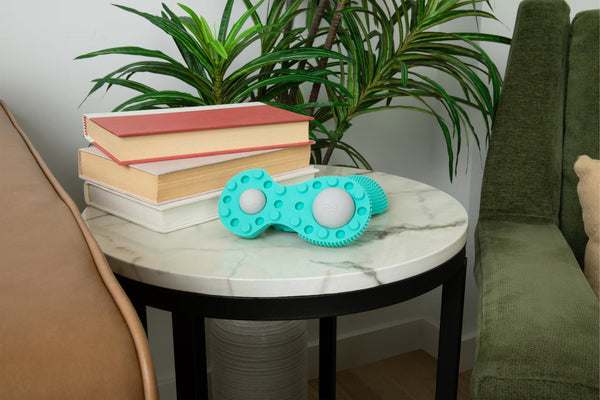 Neuro Roller | RAD wellness tool in turquoise with textured surface resting on a white marble round side table beside stacked books and green plants in a cozy living room setting.