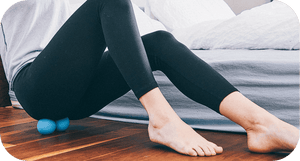 Person using a blue RAD Roller Original | RAD under their lower back on a wooden floor, demonstrating a recovery tool for muscle relief and body care.