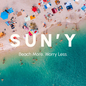 A colorful aerial view of a sandy beach with people relaxing on various towels and umbrellas near clear turquoise water, featuring the phrase "SUN'Y Beach More, Worry Less" and showcasing the Waffle Microfiber Beach Towel - Rays | SUN'Y.