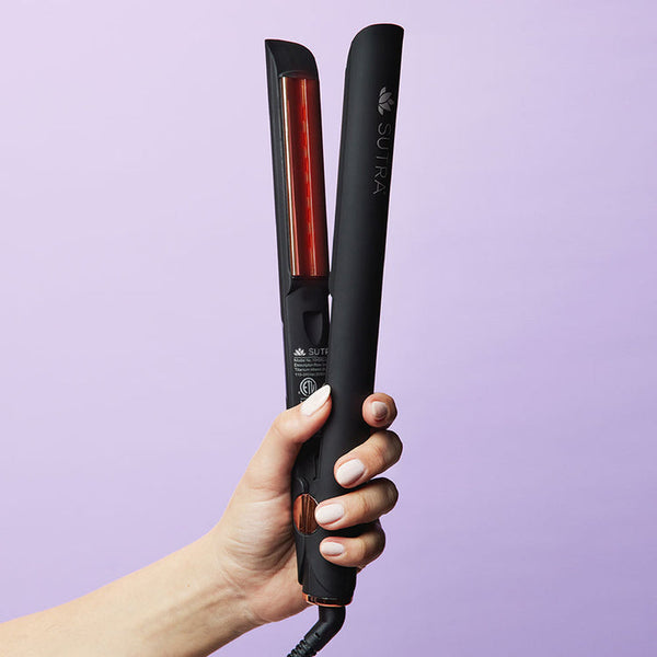 Hand holding the IR2 Infrared Flat Iron - 1" | SUTRA against a light purple background, showcasing its sleek black design and infrared heating plate.