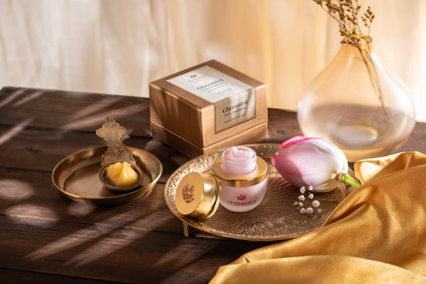 Gheesutra Face Emulsion | Shankara jar with cream on an ornate gold tray alongside its box, a brass dish with balm, a pink flower, and a translucent vase on a wooden table with warm natural light.