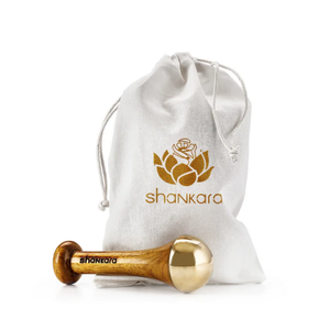 Kansa Wand Face | Shankara, a wooden and polished brass facial massage tool, pictured beside a white drawstring pouch with a gold lotus logo for skincare and wellness.