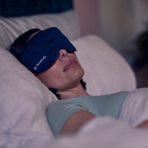 Person peacefully resting on a bed wearing a dark blue Therabody Sleepmask | Therabody designed to enhance relaxation and improve sleep quality.