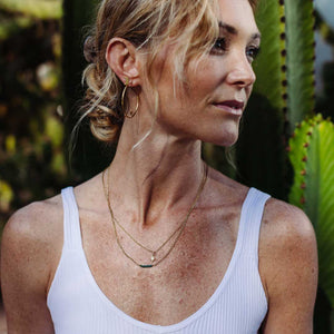 Woman wearing layered delicate gold necklaces including the North Star Necklace | Purpose Jewelry, styled with gold hoop earrings and a white tank top in an outdoor setting.