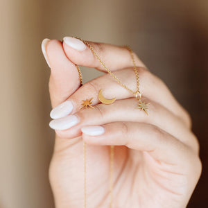 A delicate gold North Star Necklace | Purpose Jewelry held between fingers, featuring an eight-point star pendant on a fine chain against a soft background.
