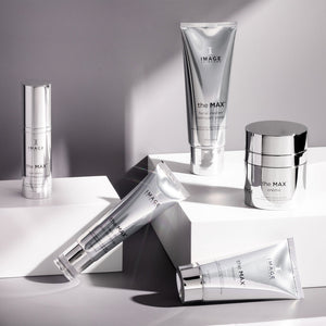 Assorted silver skincare products from the MAX™ facial cleanser | IMAGE Skincare collection arranged on white geometric platforms with soft lighting highlighting the sleek packaging.