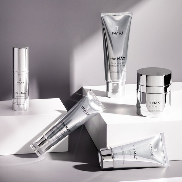 Assorted silver skincare products from the MAX™ facial cleanser | IMAGE Skincare collection arranged on white geometric platforms with soft lighting highlighting the sleek packaging.