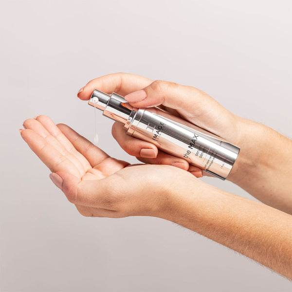 A person dispensing a clear skincare serum from a sleek silver pump bottle labeled the MAX™ serum | IMAGE Skincare into their open palm against a neutral background.
