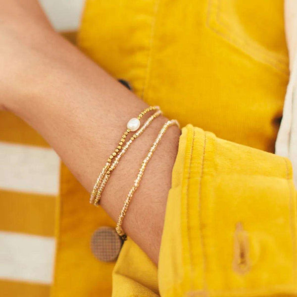 Close-up of a wrist adorned with the delicate gold Tenere Bracelet | Purpose Jewelry featuring small beads and a single white pearl, worn with a yellow jacket.