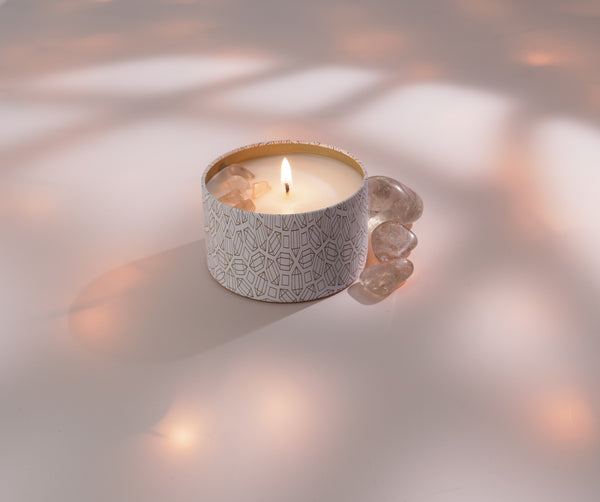 Lit candle in a decorative patterned tin with translucent stones beside it, showcasing the Defense Candle Tin 6 oz | Aluminate Life in a calming wellness setting.