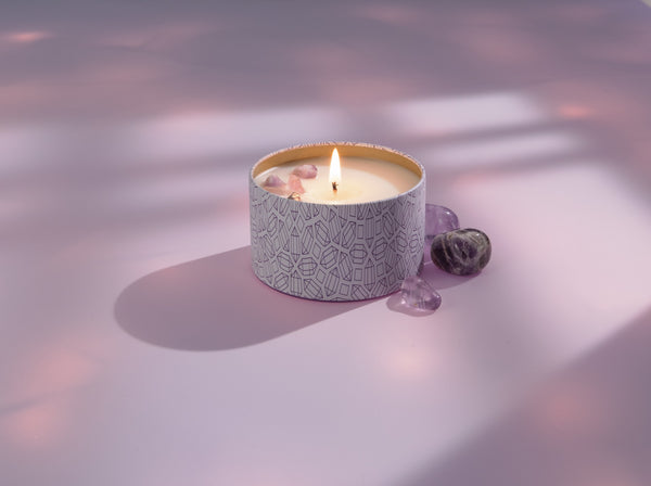 Lit Release Candle Tin 6 oz | Aluminate Life with decorative pattern, surrounded by small polished amethyst stones on a soft pink surface with diffused natural light.