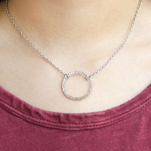 Close-up of a person wearing the Unity Necklace | Purpose Jewelry, featuring a delicate hammered silver circle pendant on a fine chain against a maroon top.