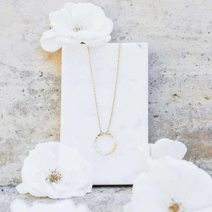 Delicate gold circular pendant necklace displayed on a white marble block with soft white flowers, showcasing the elegant Unity Necklace | Purpose Jewelry design.