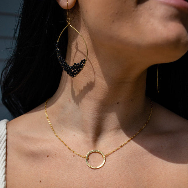 Close-up of a woman wearing the Unity Necklace | Purpose Jewelry, featuring a delicate gold-tone circular pendant on a fine chain, paired with a large elegant gold and black beaded earring.