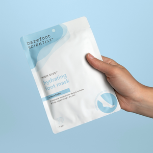 Hand holding a packet of High Dive Hydrating Foot Mask | Barefoot Scientist, designed for deep hydration and repair of rough, dry skin with shea butter.
