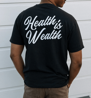 Man wearing a black Health Is Wealth T-Shirt | Lucky Owl with white cursive text on the back, standing against a white wall.