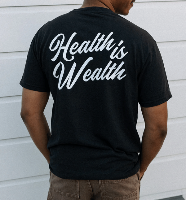 Man wearing a black Health Is Wealth T-Shirt | Lucky Owl with white cursive text on the back, standing against a white wall.