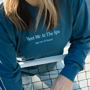 Person wearing a blue long sleeve shirt with the text "Meet Me At The Spa - Take Care Of Yourself Long Sleeve T-Shirt | Lucky Owl" leaning on a tennis net, promoting wellness and self-care.