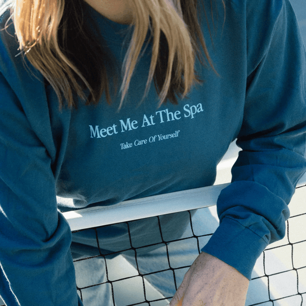 Person wearing a blue long sleeve shirt with the text "Meet Me At The Spa - Take Care Of Yourself Long Sleeve T-Shirt | Lucky Owl" leaning on a tennis net, promoting wellness and self-care.