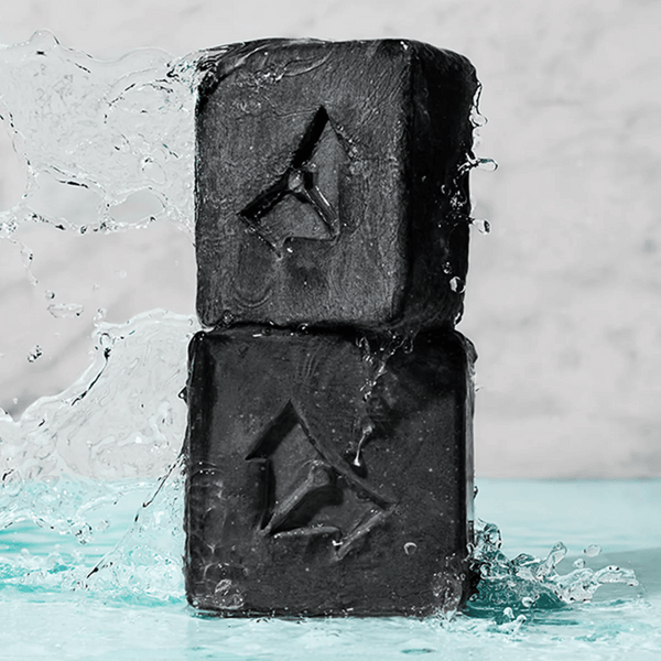 Two black charcoal cleansing bars stacked with water splashing around them, highlighting the Charcoal Cleansing Bar x Brisa Hennessy | Vertra for skincare and body care.