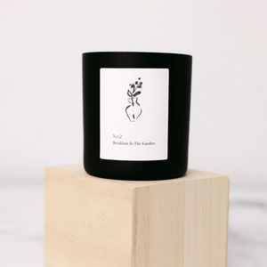 Black candle with minimalist white label featuring a simple vase illustration and text "No2 Breakfast In The Garden" displayed on a light wooden block, No. 2 Breakfast In The Garden (Swiss Alps) Candle | Lucky Owl