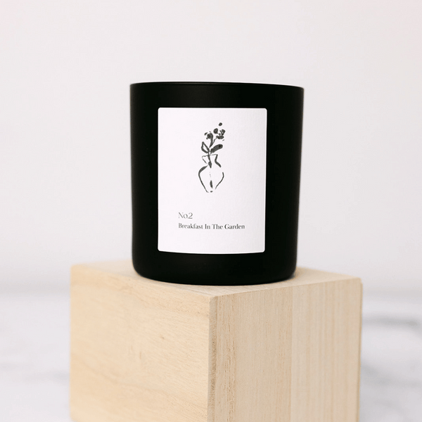 Black candle with minimalist white label featuring a simple vase illustration and text "No2 Breakfast In The Garden" displayed on a light wooden block, No. 2 Breakfast In The Garden (Swiss Alps) Candle | Lucky Owl