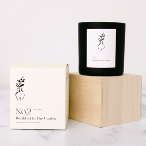 No. 2 Breakfast In The Garden (Swiss Alps) Candle | Lucky Owl in a black glass container with white minimalist label next to its cream-colored packaging box, placed on a wooden block and marble surface.