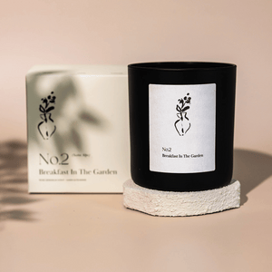 No. 2 Breakfast In The Garden (Swiss Alps) Candle | Lucky Owl in a black glass container with minimalist white label featuring a floral illustration, placed on a textured white stone coaster with matching product box in the background.