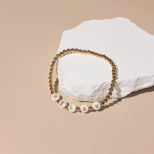 Close-up of a gold beaded bracelet with white beads spelling "RELAX," part of the Wooden Beaded + Gold Bead 3 Bracelet Set - Relax | Lucky Owl, displayed on a white textured stone against a beige background.