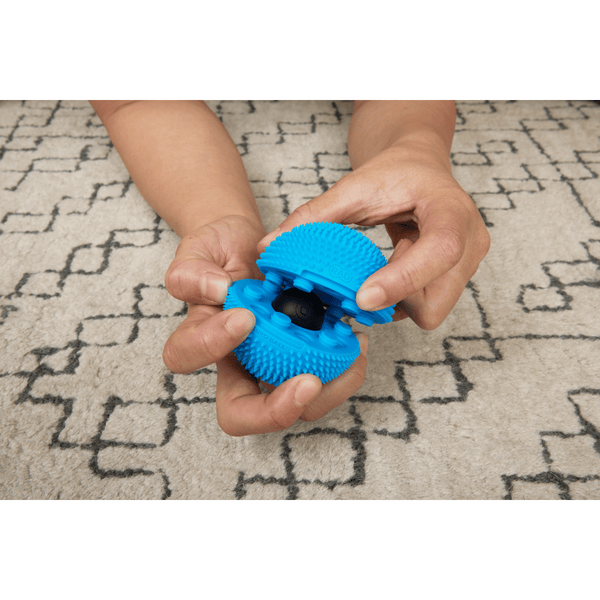 Hands holding and opening a bright blue textured Neuro Ball | RAD wellness device designed for muscle recovery and body care on a patterned carpet background.