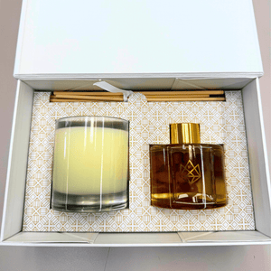 Open gift box containing a Candle & Diffuser Set - Cheer | Aluminate Life with a yellow candle in a glass holder, amber diffuser bottle with gold cap, and several diffuser sticks on a patterned base.