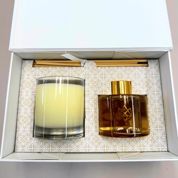 Open gift box containing a Candle & Diffuser Set - Cheer | Aluminate Life with a yellow candle in a glass holder, amber diffuser bottle with gold cap, and several diffuser sticks on a patterned base.