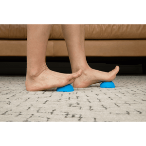 Person using blue textured Neuro Ball | RAD devices under bare feet on a carpeted floor, demonstrating a foot massage and recovery tool for wellness and body care.