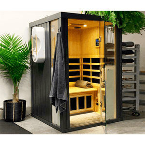 Modern black and wood HaloSauna™ | Halotherapy Solutions unit with glass door, interior seating, digital control panel, and gray towel hanging outside, placed next to a green plant for wellness and skincare.