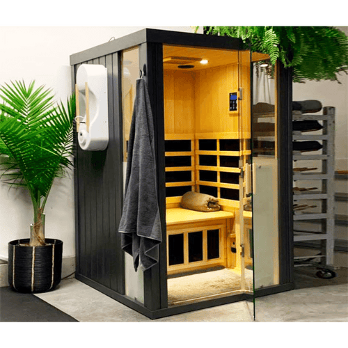 Modern black and wood HaloSauna™ | Halotherapy Solutions unit with glass door, interior seating, digital control panel, and gray towel hanging outside, placed next to a green plant for wellness and skincare.
