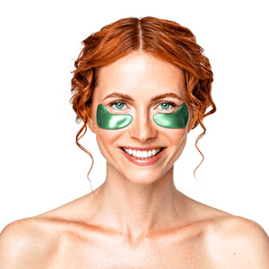 Smiling woman with red hair wearing Green Jade Eye Mask | KNESKO under her eyes, promoting skincare and wellness recovery tools.