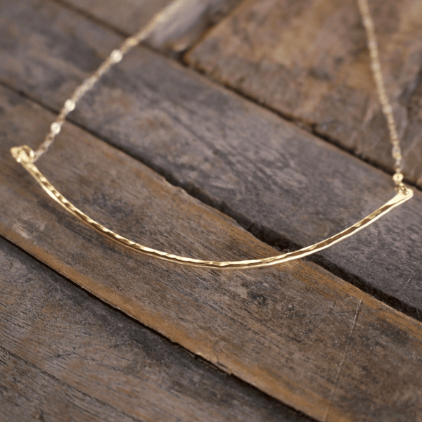 Close-up of a textured curved gold bar necklace pendant on a delicate chain, styled on a rustic wooden surface, showcasing the Rounded 14K Gold Fill Bar Necklace | Liv & B.