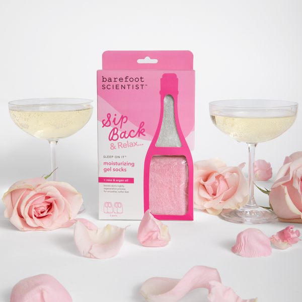 Sip Back & Relax | Barefoot Scientist moisturizing gel socks with rose and argan oil displayed with pink roses, petals, and two glasses of sparkling drink suggesting a soothing self-care moment.