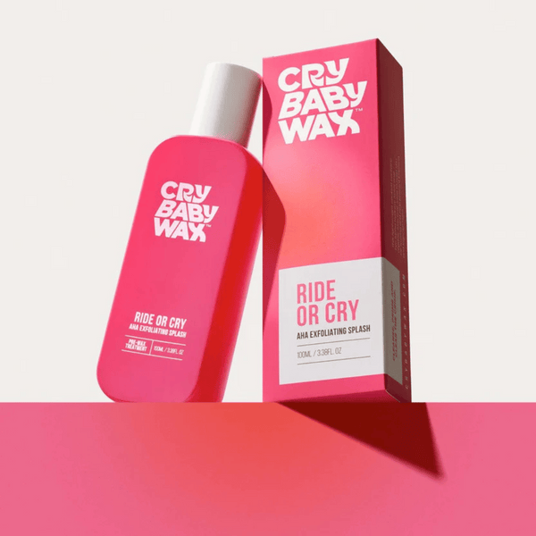 Bright pink bottle and matching box of Ride or Cry AHA Exfoliating Splash by Crybaby Wax displayed on a pink surface with a light background, highlighting skincare product packaging.