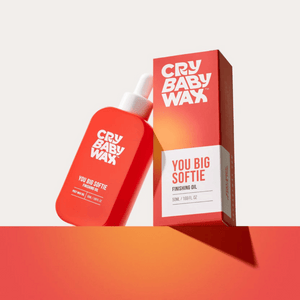 Red bottle and matching box packaging of You Big Softie Finishing Oil | Crybaby Wax with a dropper cap, displayed on an orange surface against a light background.