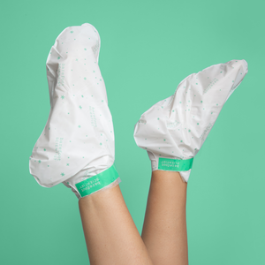 Legs wearing white moisturizing foot masks with green accents against a green background, showcasing the High Dive Hydrating Foot Mask | Barefoot Scientist for skincare and body care.