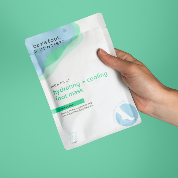 Hand holding a package of High Dive Hydrating Foot Mask | Barefoot Scientist against a teal background, featuring cooling eucalyptus ingredients for skincare and wellness.