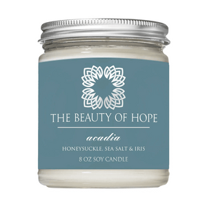 Acadia 8oz Candle | The Beauty of Hope featuring a soy wax candle in a clear glass jar with a silver lid and a blue label highlighting honeysuckle, sea salt, and iris scent.