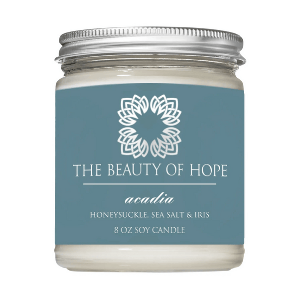 Acadia 8oz Candle | The Beauty of Hope featuring a soy wax candle in a clear glass jar with a silver lid and a blue label highlighting honeysuckle, sea salt, and iris scent.