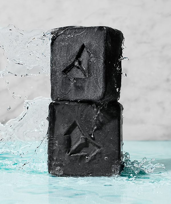Two black charcoal cleansing bars stacked with water splashing around them on a light blue surface, featuring the product name Charcoal Cleansing Bar x Brisa Hennessy | Vertra.