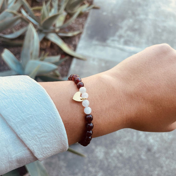 Close-up of a wrist wearing a Gemstone Heart Bracelet | Purpose Jewelry featuring dark red and white beads with a small gold heart charm, outdoors near succulent plants.